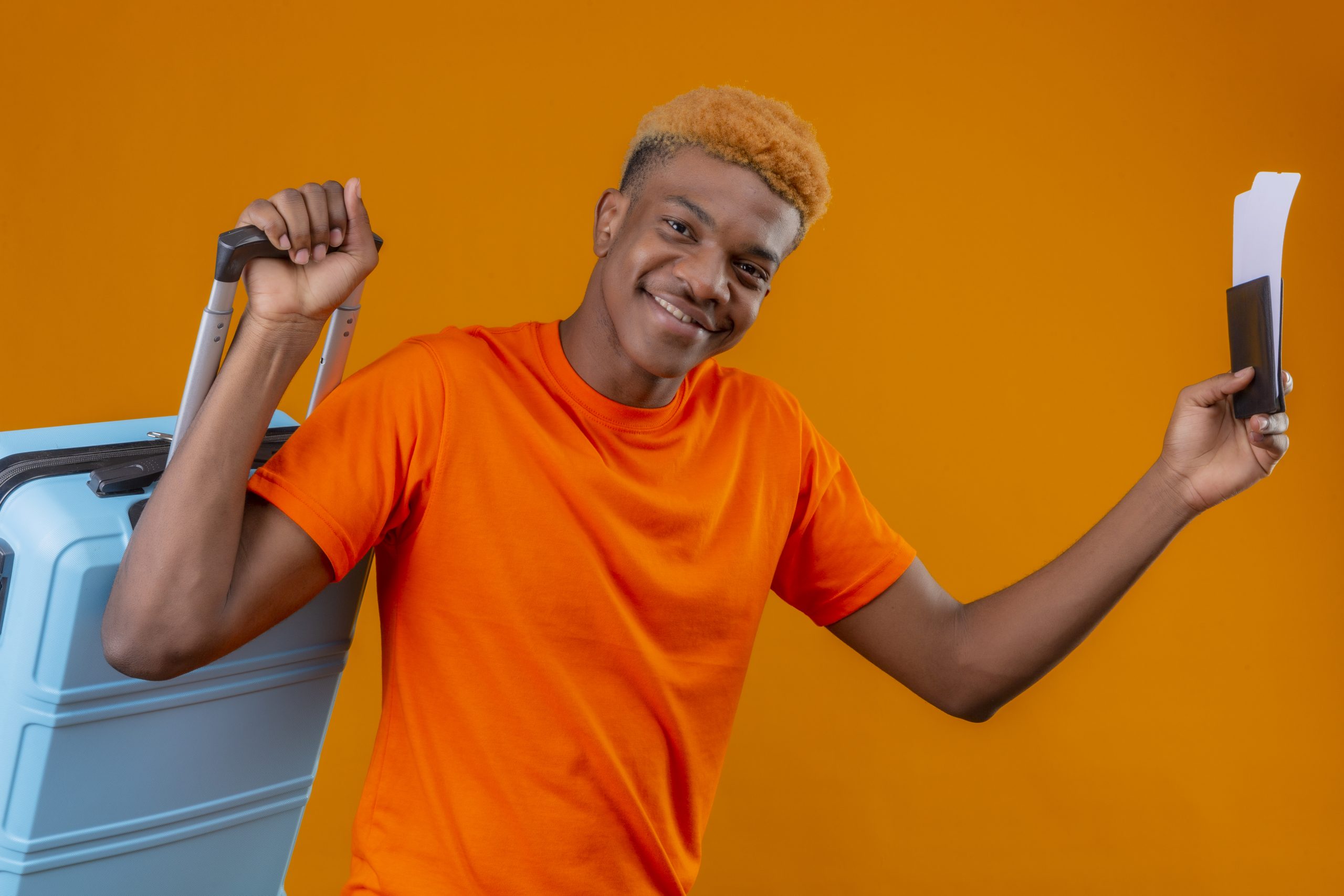 pleased young african american handsome boy wearing orange t-shirt holding travel suitcase and air tickets smiling happy and positive standing over orange background